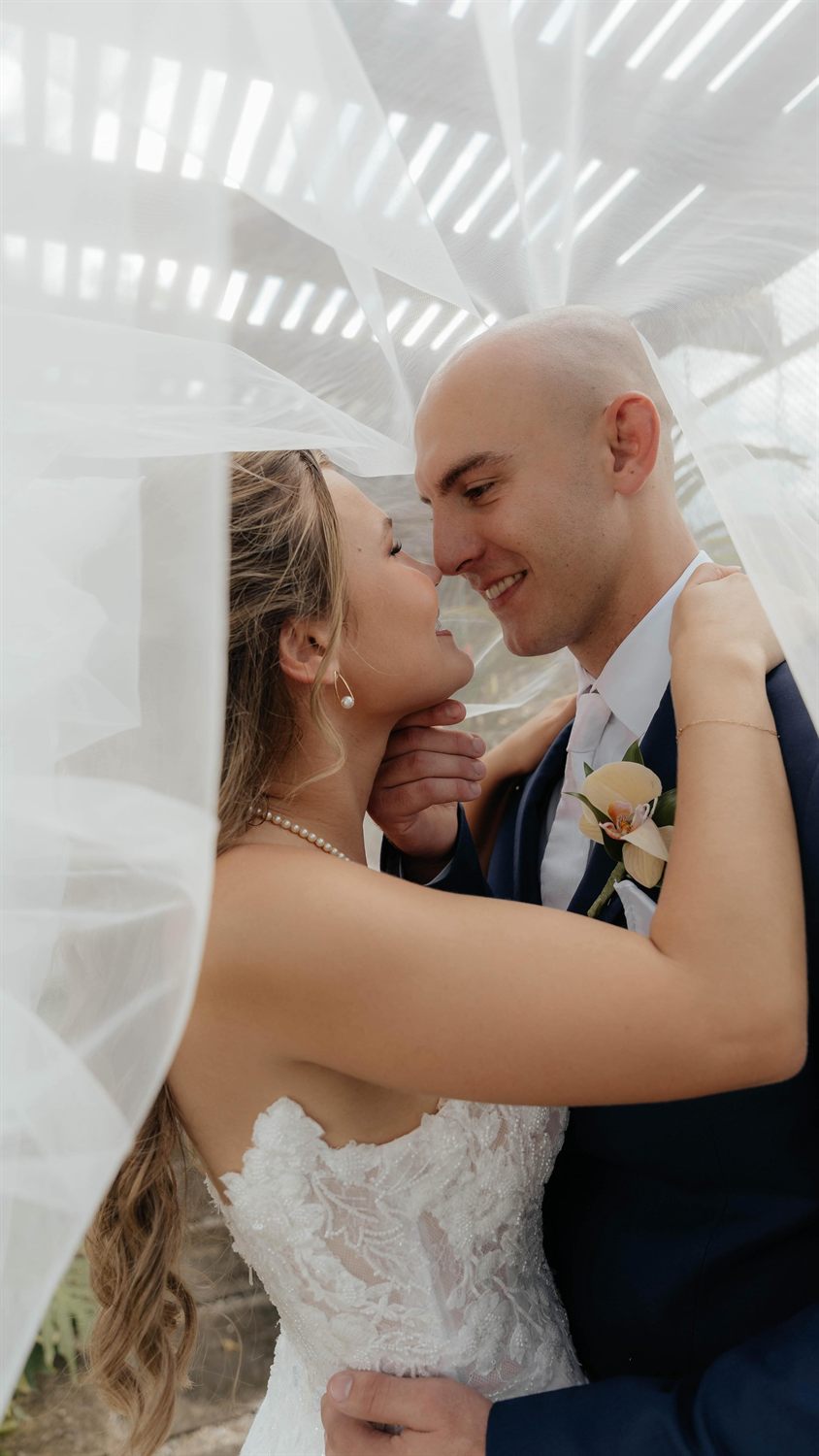 A bride and groom smile beneath a flowing veil