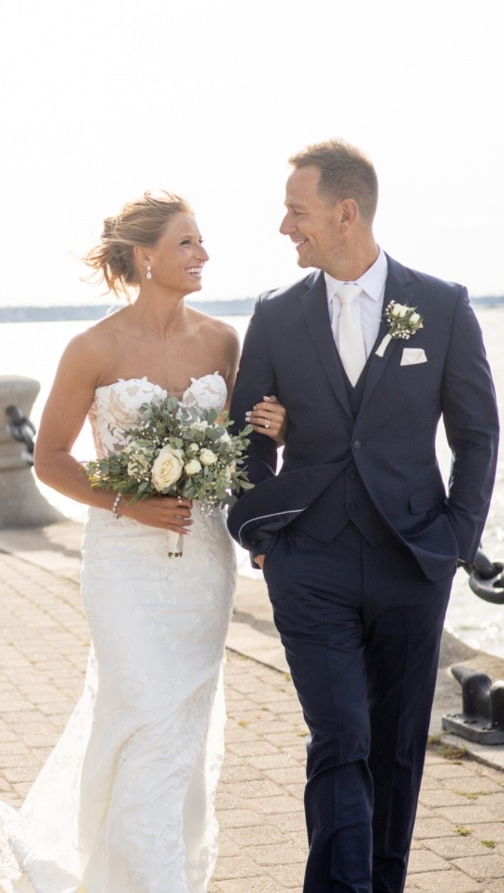 Lexi and John with their wedding party walking together beneath a floral arch on their wedding day