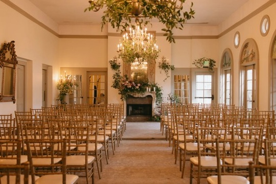 An elegant ceremony space lined with gold chiavari chairs leading to a floral-draped fireplace